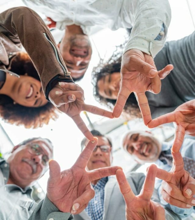 multiracial group of formally dressed coworkers between the ages of 25 and 50 are inside the company they work for and are photographed as they put their heads together and look down at the camera that portrays them